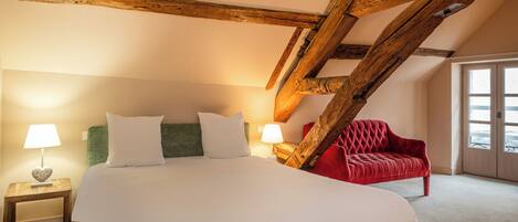 Une chambre avec un grand lit, deux tables de chevet avec des lampes, un fauteuil rouge et des poutres apparentes.