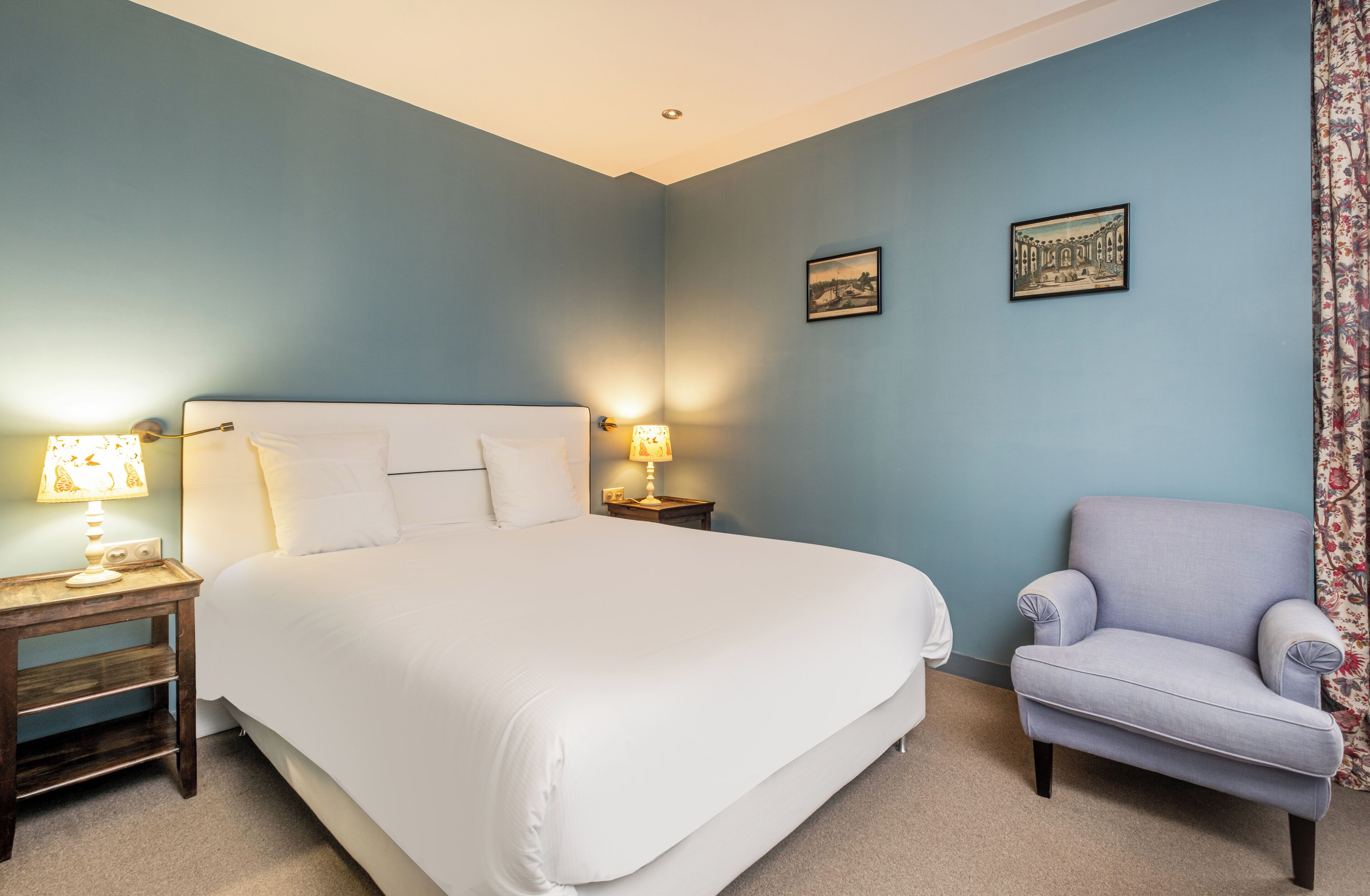 A bedroom with a large bed, two bedside tables with lamps, a chair, and two framed pictures on the wall.