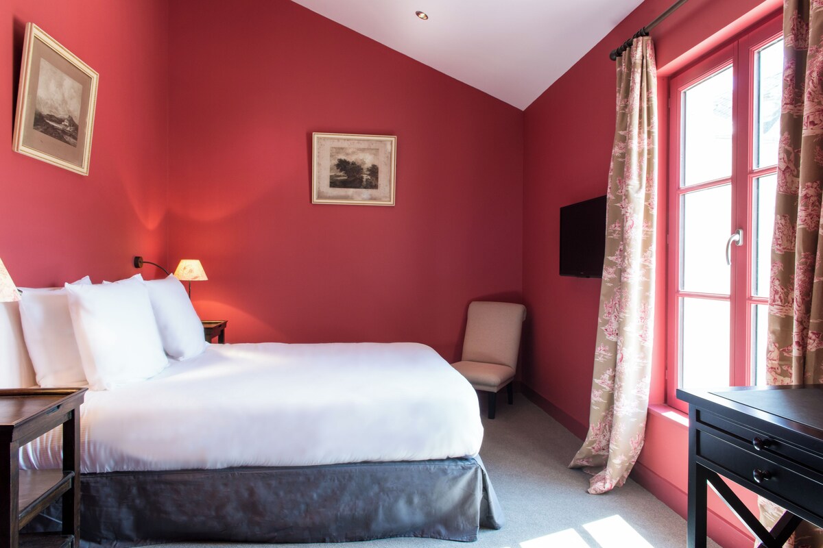 Quarto de hotel com cama, uma cadeira, escrivaninha e janela com cortinas.