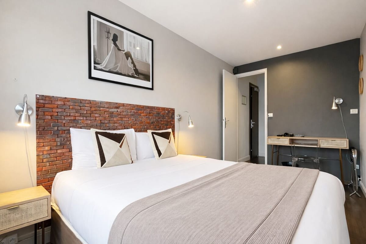 A modern bedroom with a brick headboard, a large bed, bedside lamps, a desk, and a framed picture on the wall.