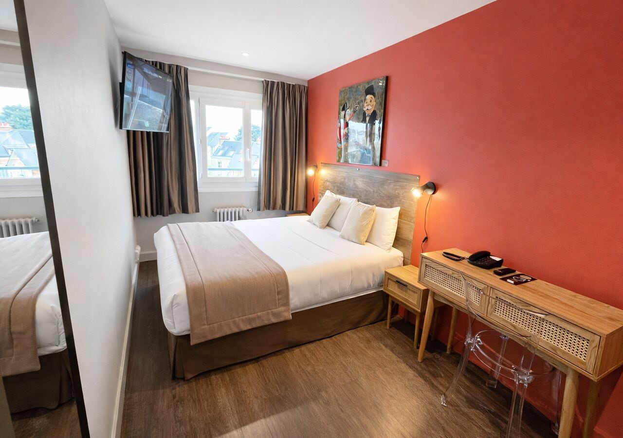 A hotel room with a bed, a desk with a chair, a TV mounted on the wall, and a window with curtains.