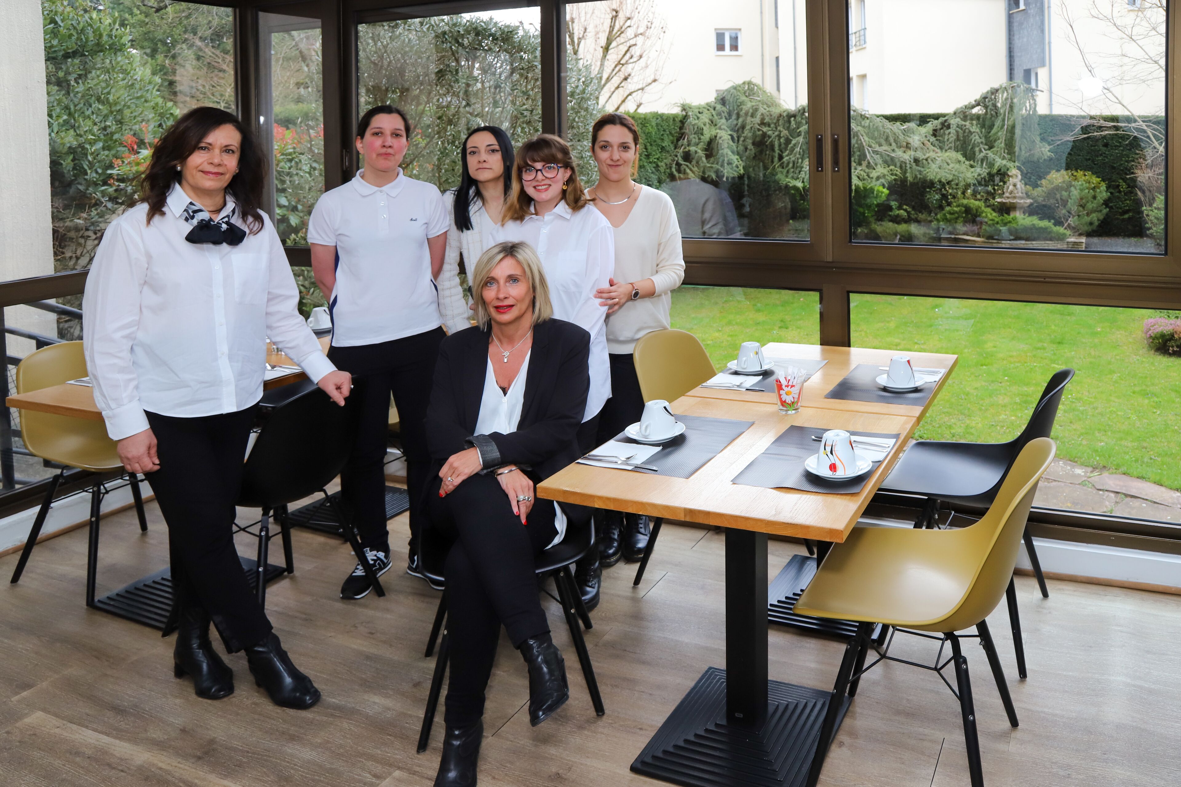 Un grupo de personas sentadas y de pie en un espacio interior moderno con ventanas amplias, una mesa de madera y sillas amarillas.