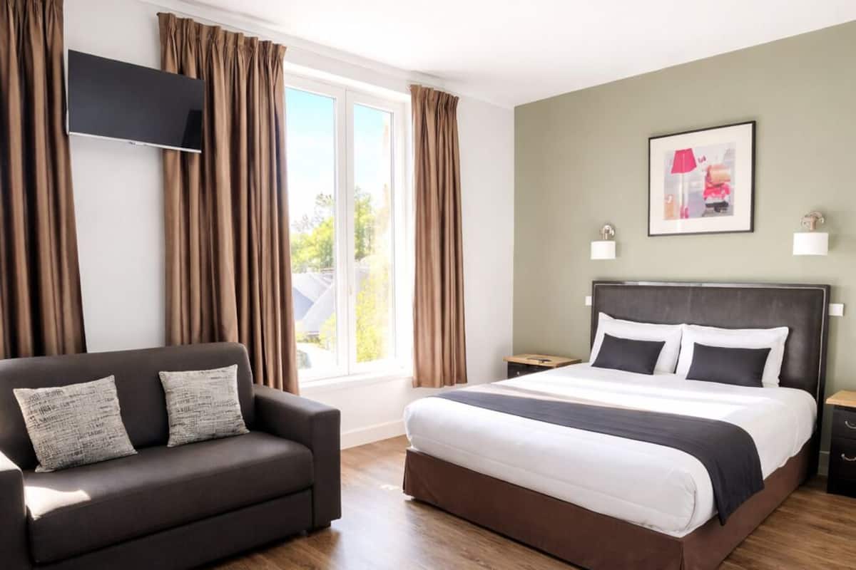 A modern hotel room with a bed, a sofa, a flat-screen TV, and a window with curtains.