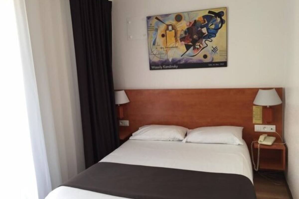 Una habitación de hotel con una cama grande, dos lámparas de noche, una mesita con un teléfono y un cuadro en la pared.