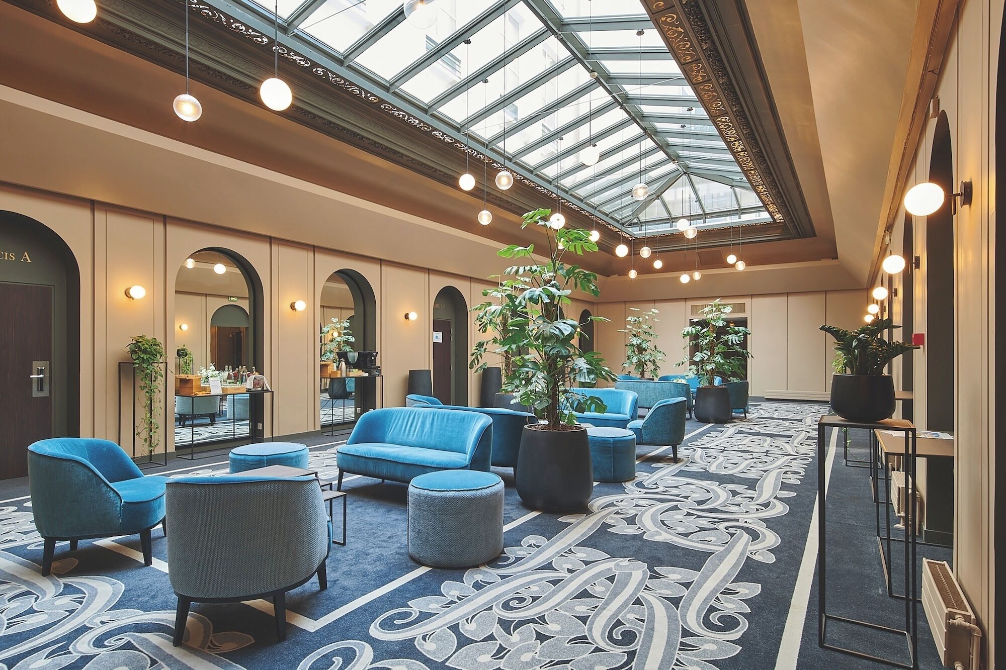 A hotel lobby with a high ceiling, skylight, blue seating, potted plants, and a patterned carpet.