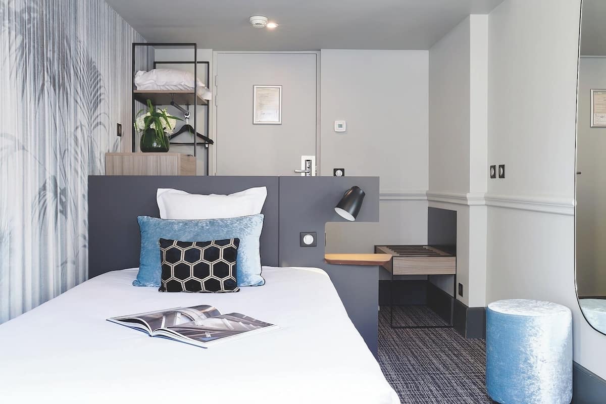 A modern hotel room with a bed, a desk, a chair, a lamp, a plant, and a waste bin.
