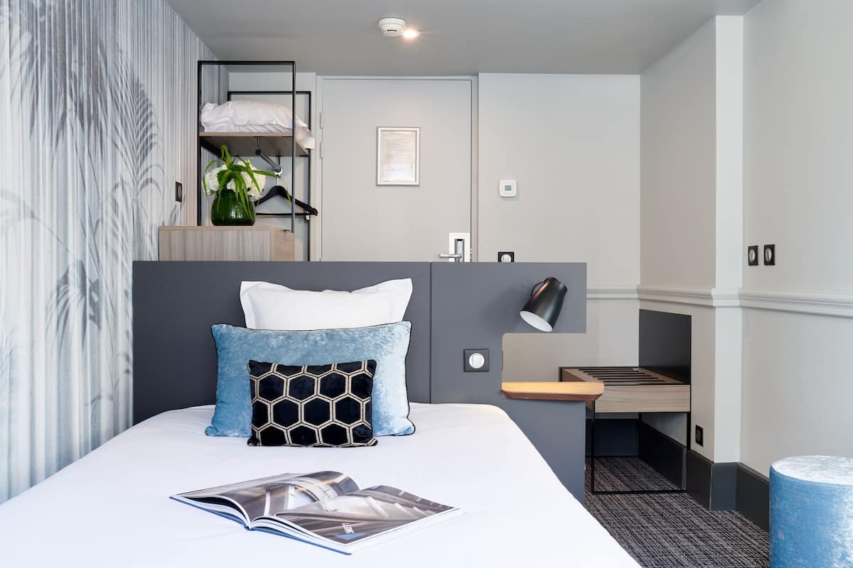 A modern hotel room with a neatly made bed, a small desk with a lamp, and a compact kitchenette.
