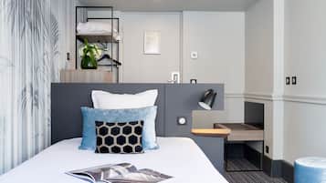 A modern hotel room with a neatly made bed, a small desk with a lamp, and a compact kitchenette.