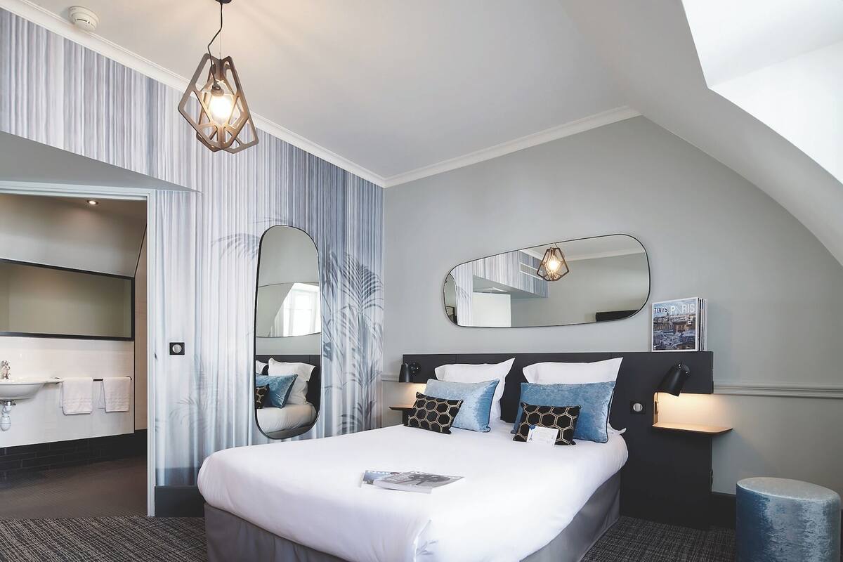 A modern hotel room with a large bed, a bathroom with a sink, and a mirror.