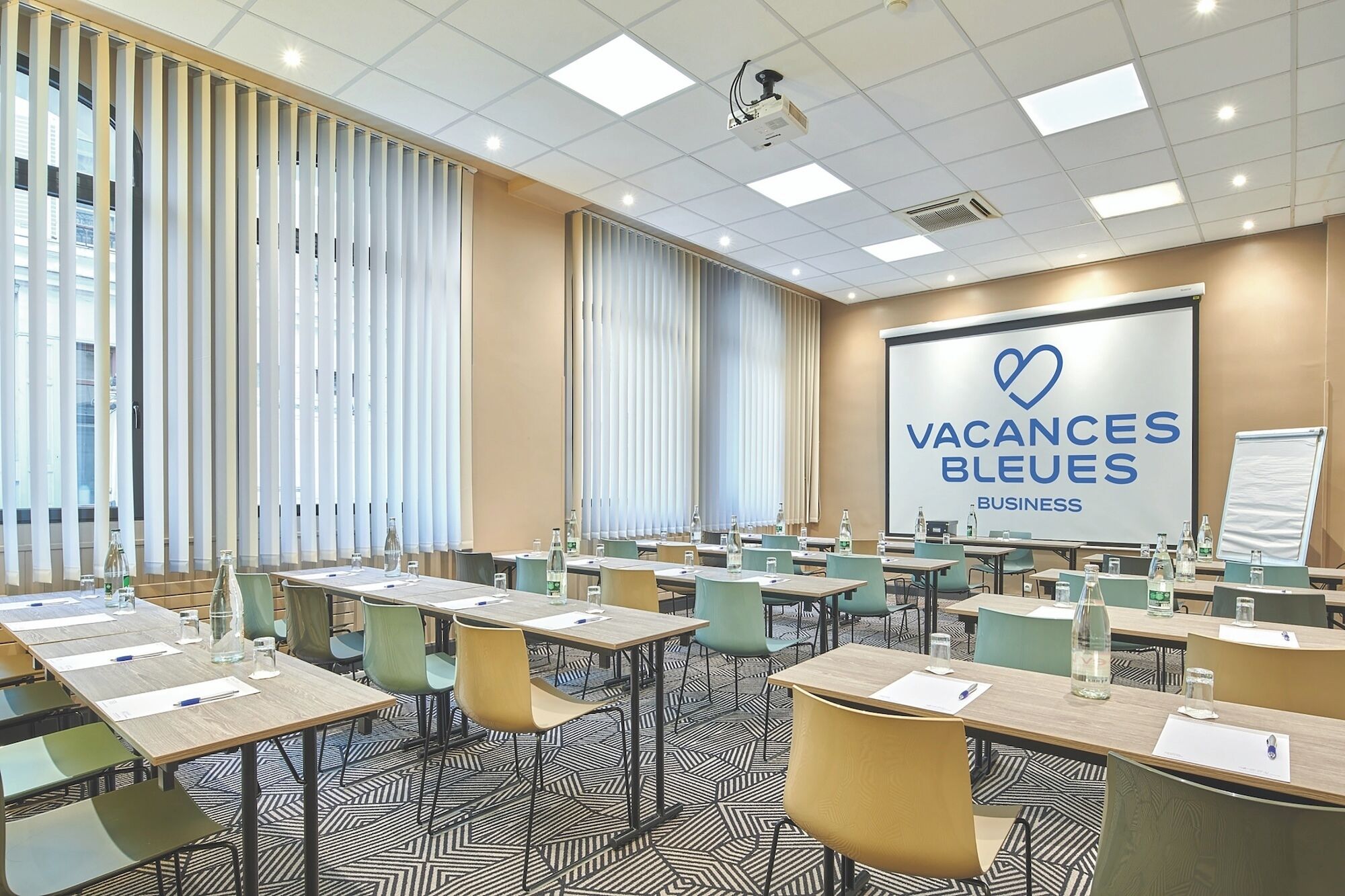 A conference room with a projector screen displaying 'Vacances Bleues Business,' tables with chairs, and a patterned carpet.