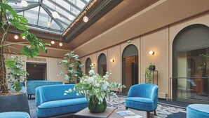 Vestíbulo de hotel con techo alto, asientos azules, una mesa central con un jarrón de flores y un atrio de cristal.