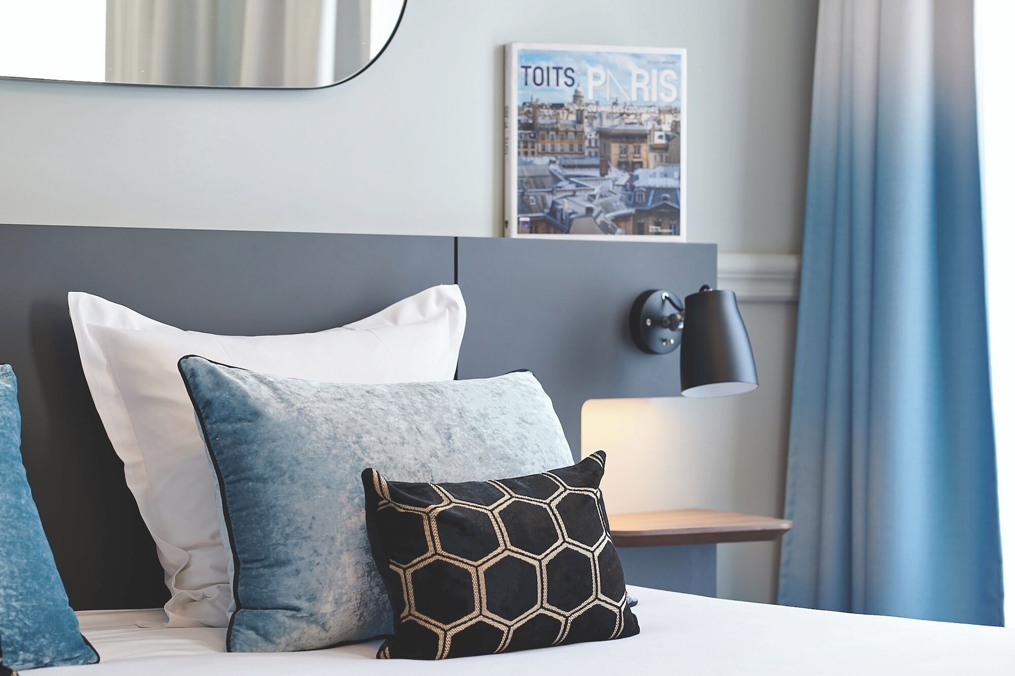 A neatly made bed with white and blue pillows, a black geometric pillow, a wall-mounted lamp, and a framed picture of Paris.