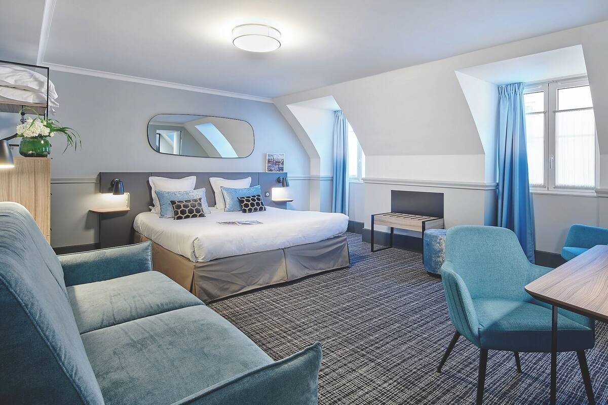 A hotel room with a bed, two armchairs, a small table, a desk, a mirror, a lamp, and a window with curtains.