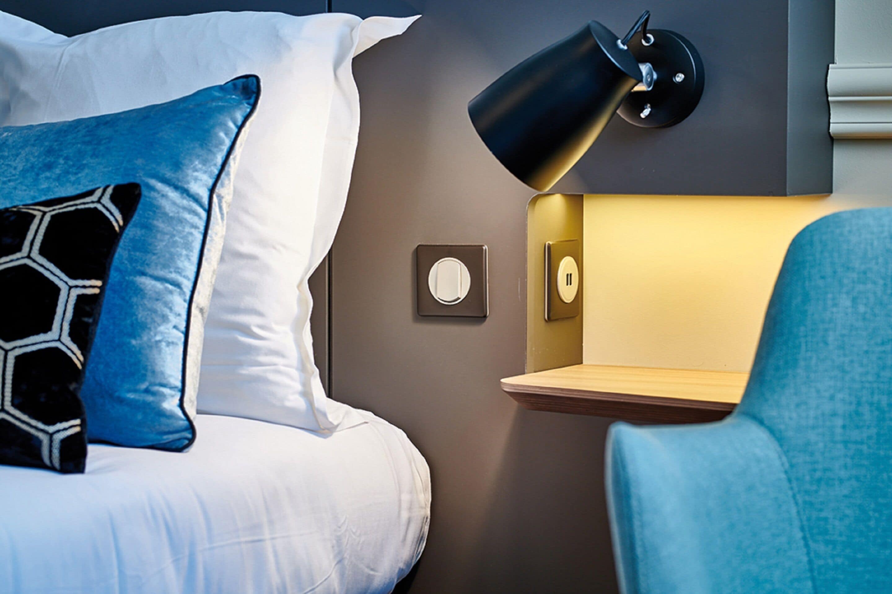 A neatly made bed with white and blue pillows, a wall-mounted lamp, and a small wooden shelf.