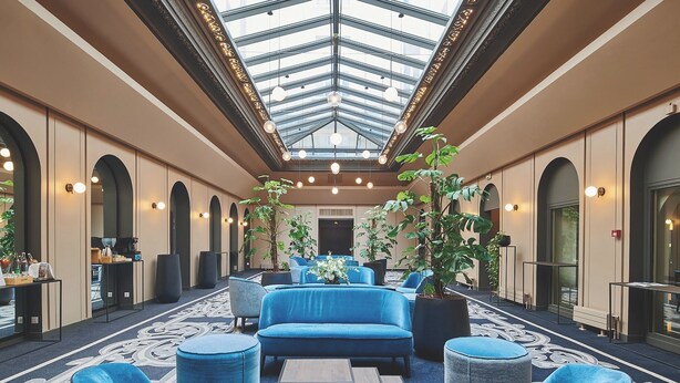 Un vestíbulo de hotel con una zona de estar central, techo de cristal y plantas en macetas.