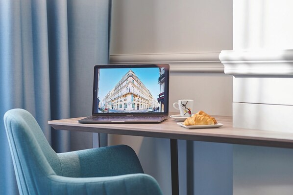 A laptop on a table with a croissant and a cup, a blue chair, and a window with curtains.