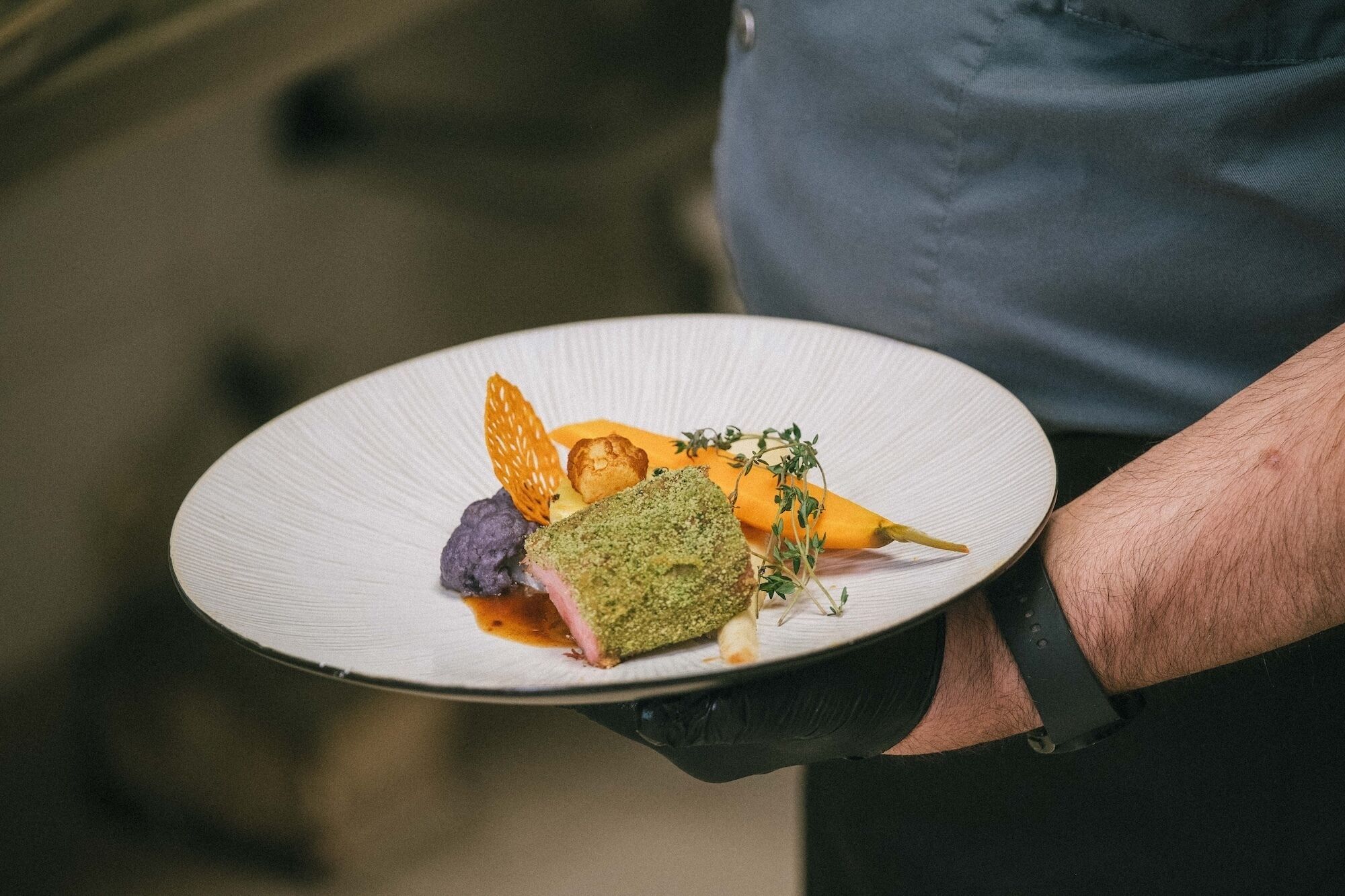A person holding a white plate with a dish that includes a green herb crust, purple element, and orange garnish.