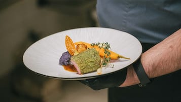 A person holding a white plate with a dish that includes a green herb crust, purple element, and orange garnish.