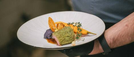A person holding a white plate with a dish that includes a green herb crust, purple element, and orange garnish.