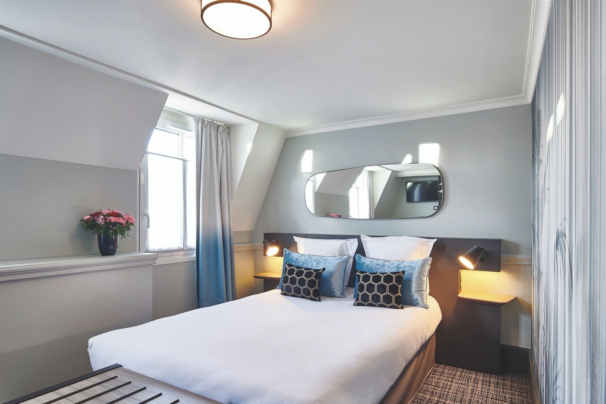 A hotel room with a bed, a mirror, a bedside table with a lamp, a window with curtains, and a vase with flowers.