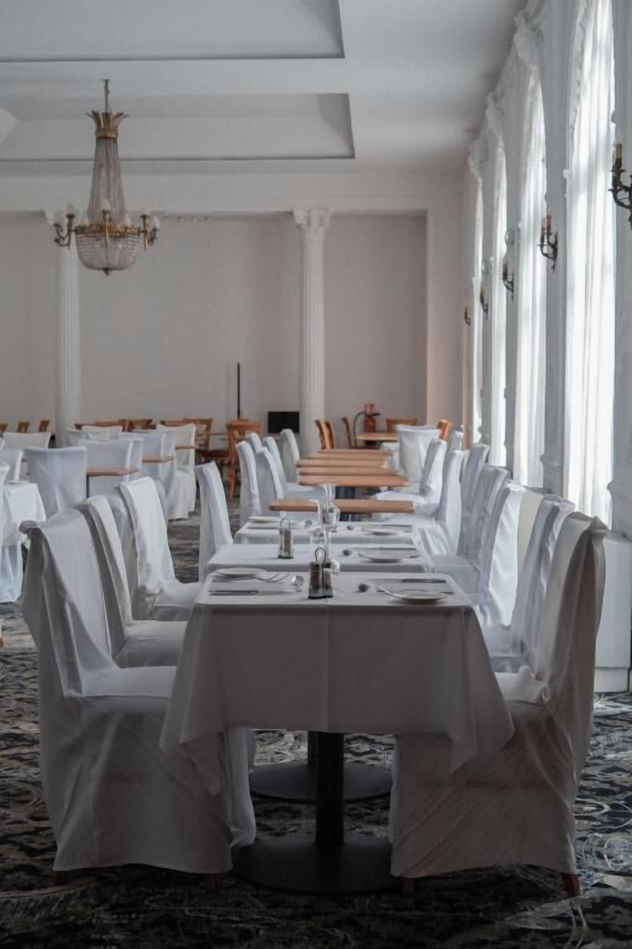 A banquet hall with long tables set for dining, white drapery on chairs, and a chandelier.