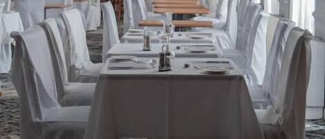 A banquet hall with long tables set for dining, white drapery on chairs, and a chandelier.