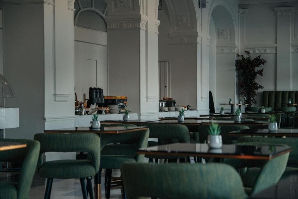 Un restaurante con sillas y mesas tapizadas de verde, columnas blancas y una zona de bar con equipo de cocina.