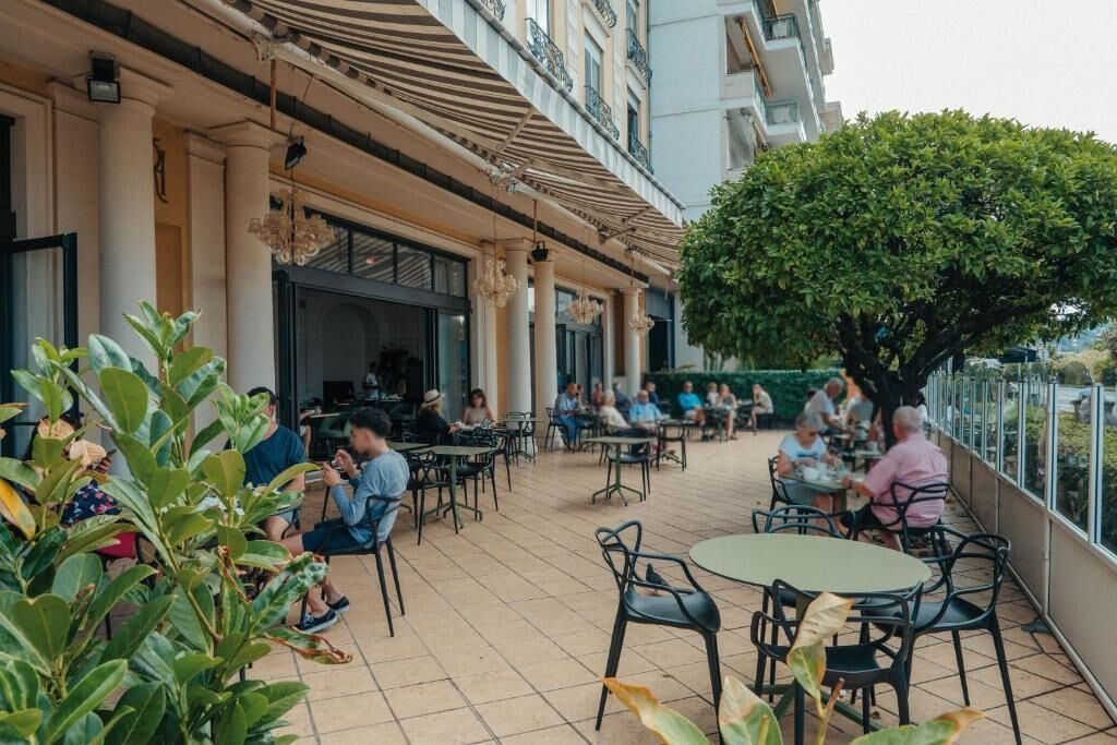 Un espace de café en plein air, avec des tables et des chaises, entouré de verdure et bordé par la façade d’un bâtiment.