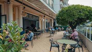 Un espace de café en plein air, avec des tables et des chaises, entouré de verdure et bordé par la façade d’un bâtiment.