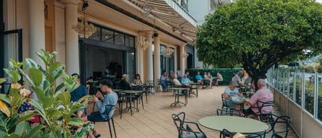 An outdoor cafe area with tables and chairs, surrounded by greenery and a building facade.