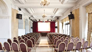 A conference room with rows of chairs, a stage with a red curtain, and a chandelier.