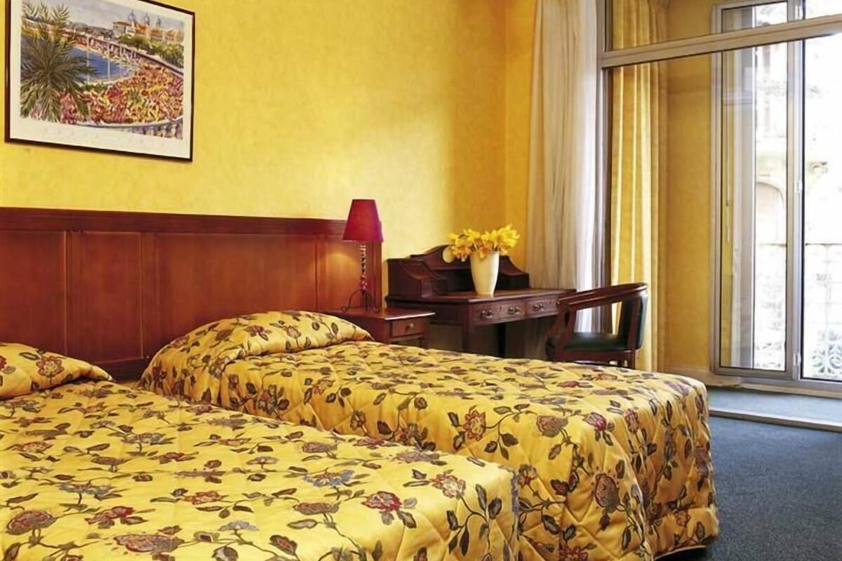 A hotel room with two beds, a wooden headboard, a desk with a lamp, a vase with flowers, and a window with curtains.