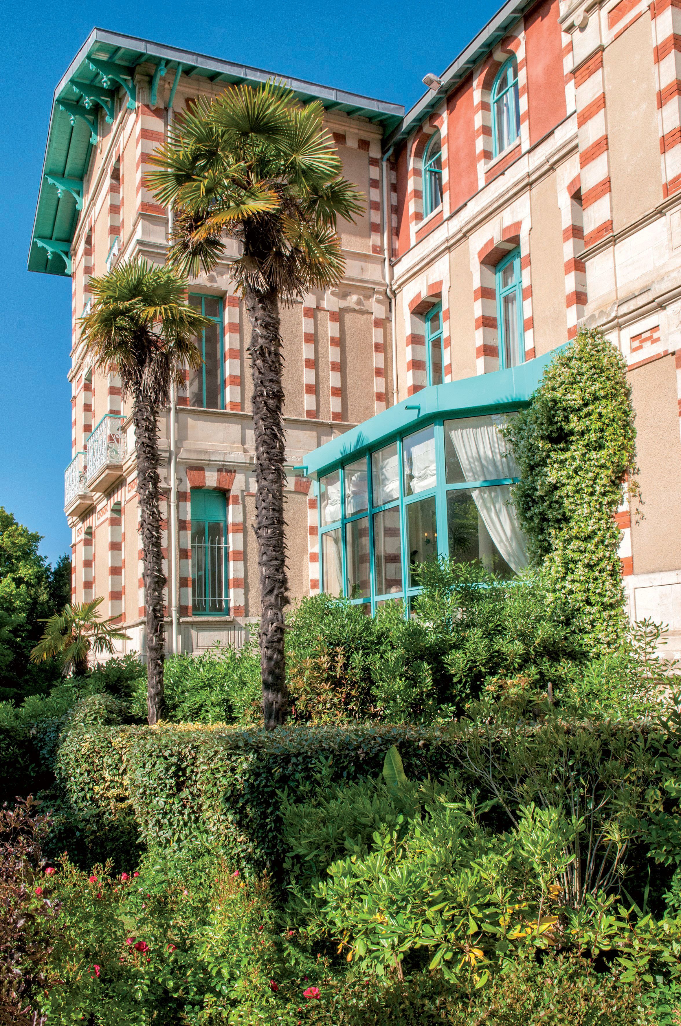 Un edificio histórico con fachada a rayas, ventanas grandes y un jardín exuberante con palmeras y flores rojas.