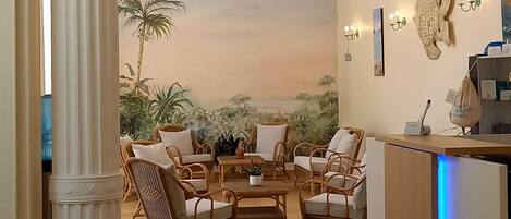 A room with a wooden herringbone floor, a reception desk with a blue light, wicker furniture, a large column, and a mural of a tropical landscape.