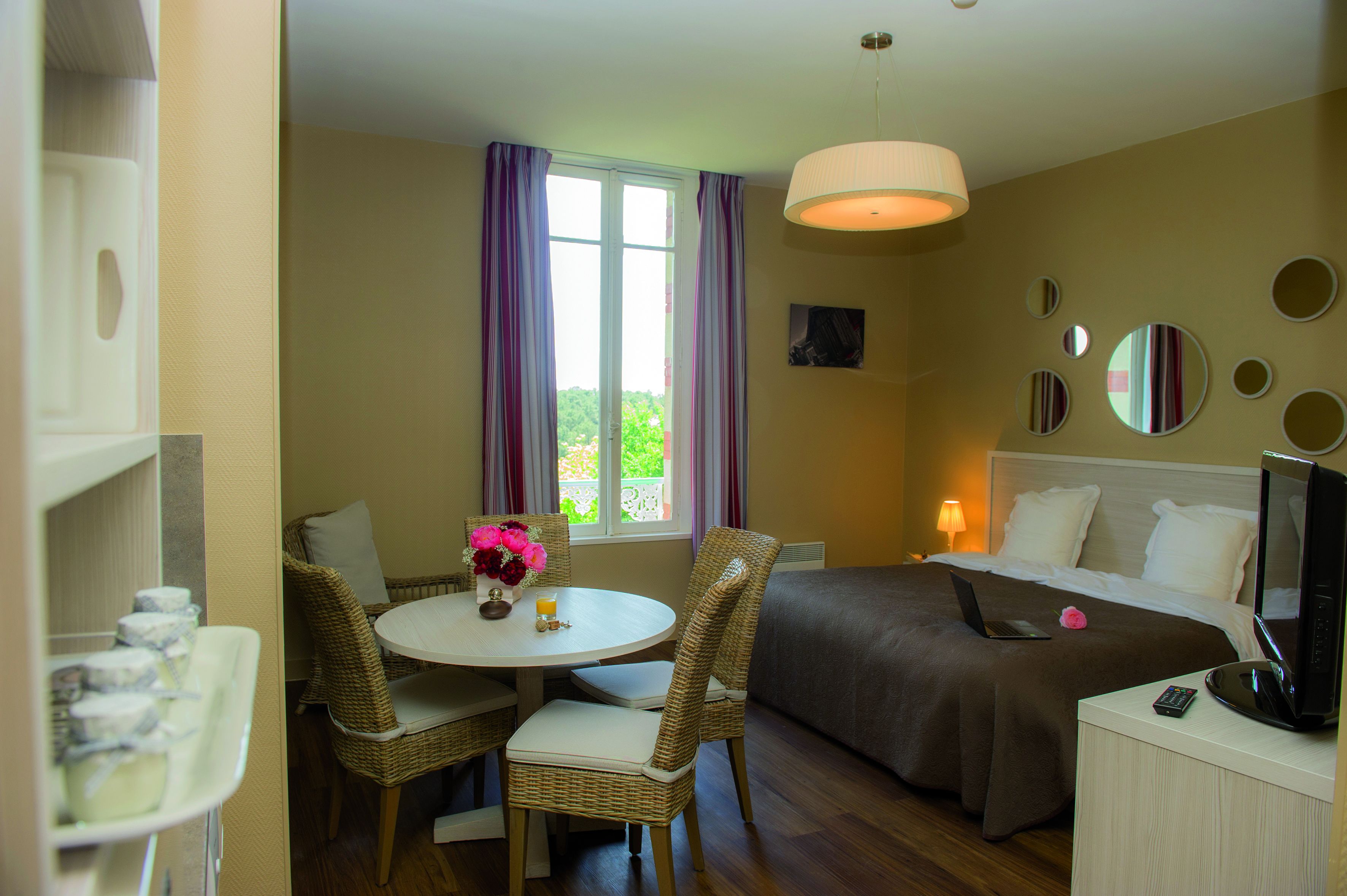 Habitación de hotel con una cama, un escritorio con una computadora portátil, una mesa de comedor con sillas y una ventana con cortinas.