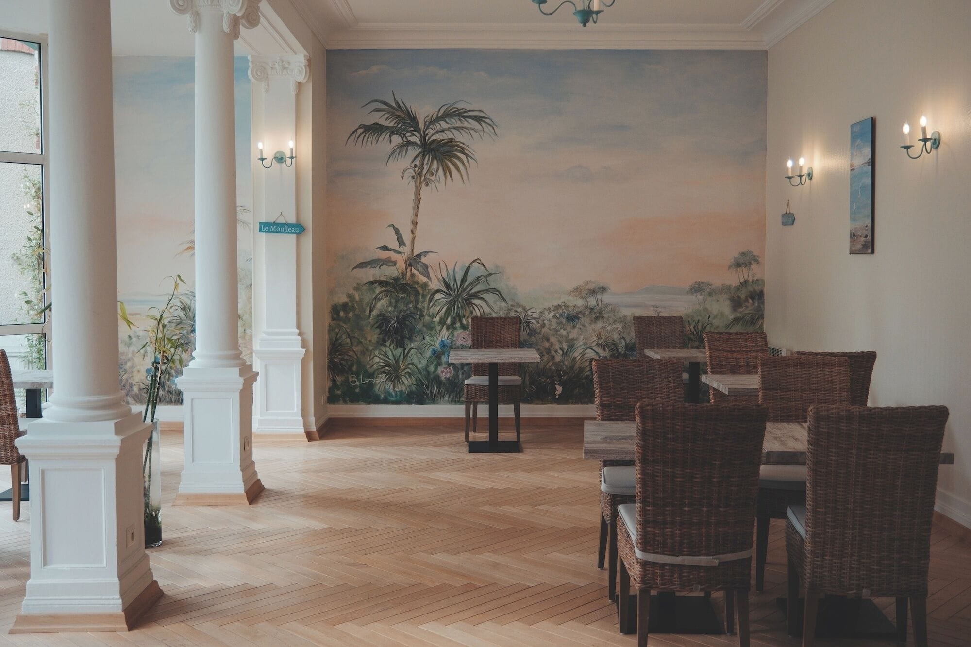 Un coin repas avec des chaises en osier, une fresque représentant un paysage tropical et une enseigne indiquant « Le Mouilleux ».