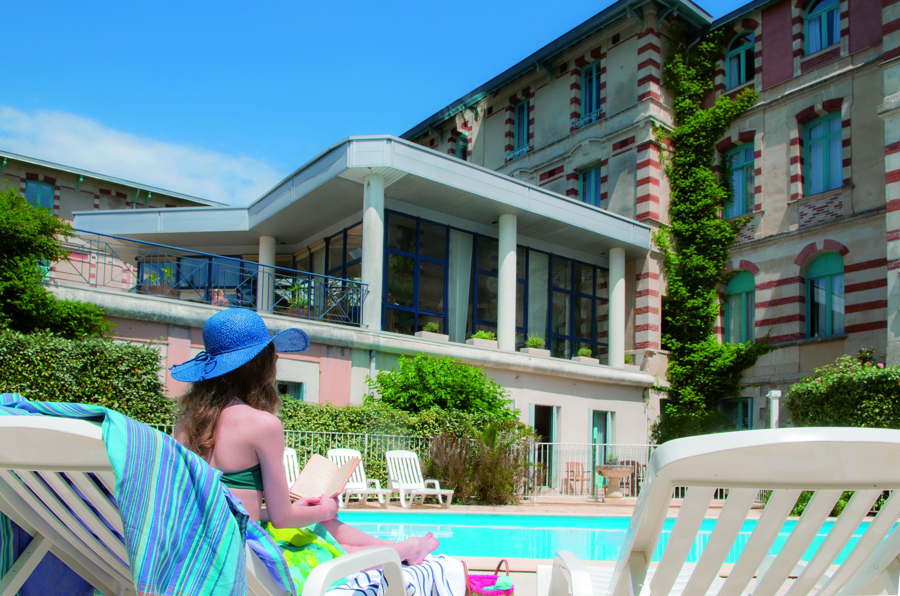 Un espace aménagé au bord de la piscine, avec des chaises longues, et un bâtiment doté de grandes fenêtres et d’un balcon.