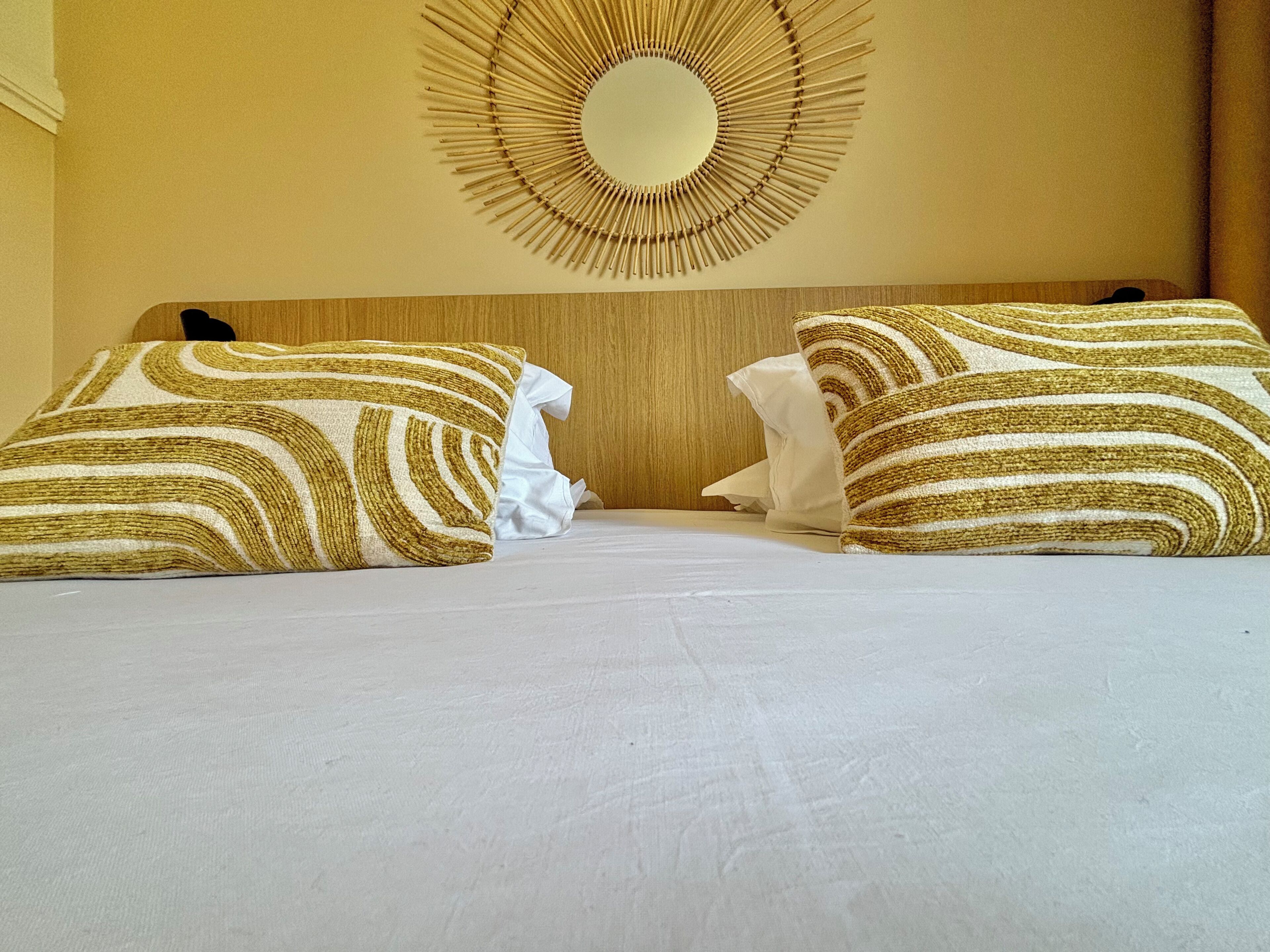 A hotel room with a bed, two pillows with a striped pattern, a round mirror on the wall, and a wooden headboard.