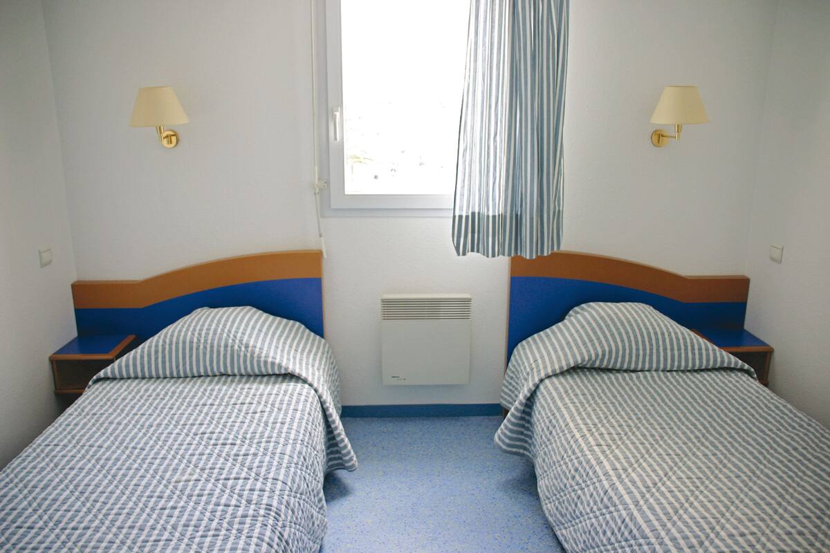 A room with two single beds, each with a headboard and a bedside table. There are wall-mounted lamps on either side of a window with a curtain.