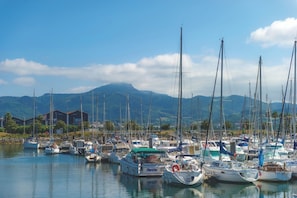 A marina with multiple boats docked, a mountain in the background, and a building with a red roof. - Hôtel Résidence Vacances Bleues Orhoitza (Hendaye)