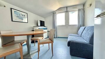 A compact room with a wooden dining table, two chairs, a sofa, a television on a cabinet, and a window with curtains.