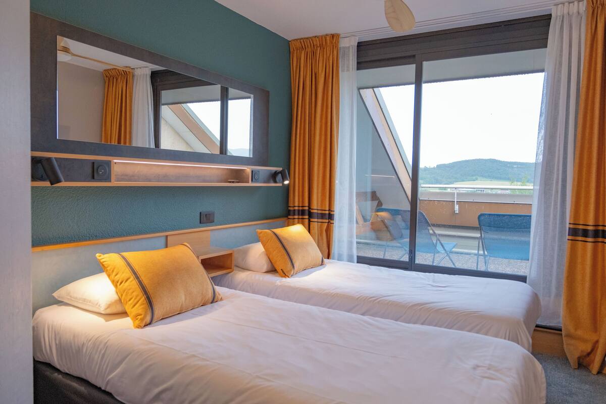 Een hotelkamer met twee bedden, een groot raam met gordijnen, een balkon met stoelen en uitzicht op het landschap.