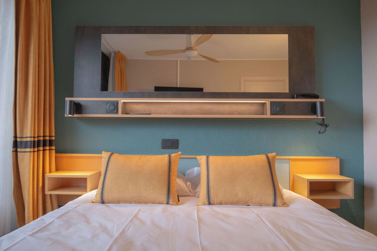 Een opgemaakt bed met twee kussens, een nachtkastje, een wandplank met spiegel en een plafondventilator.