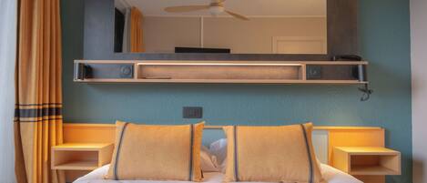 A neatly made bed with two pillows, a bedside table, a wall-mounted shelf with a mirror, and a ceiling fan.