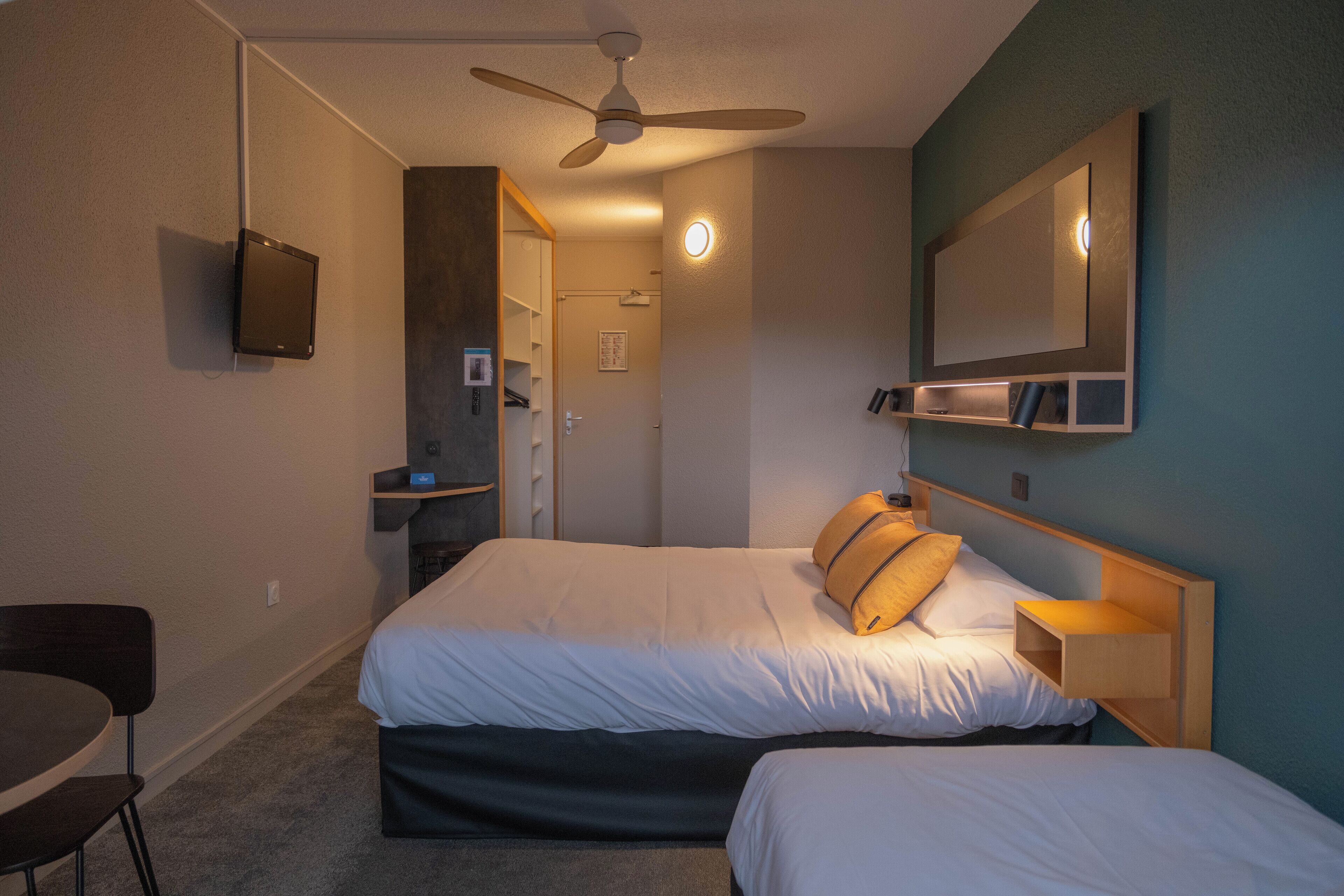 A hotel room with a bed, a desk, a chair, a ceiling fan, a TV, and a mirror.