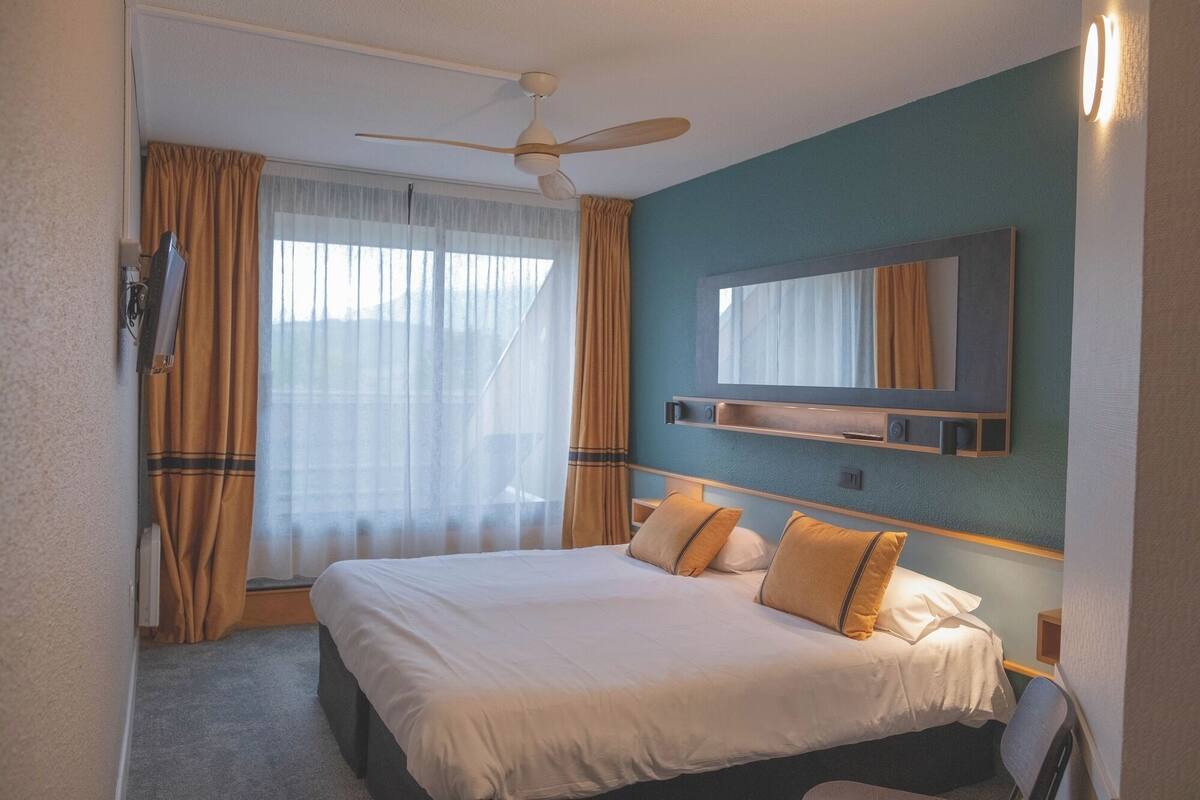 Hotelkamer met een bed, plafondventilator, groot raam met gordijnen, een aan de muur bevestigde televisie en een nachtkastje.