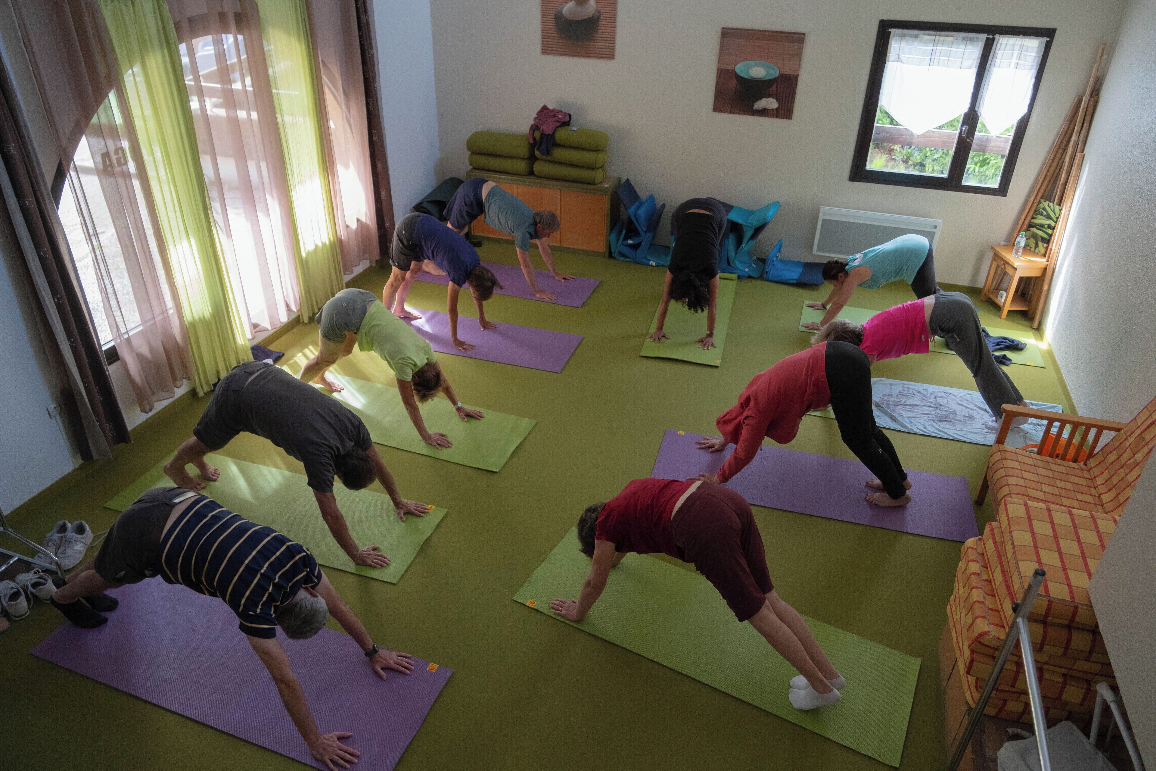 A group of people doing yoga on mats in a room with a green floor, a window, and a chair in the background.