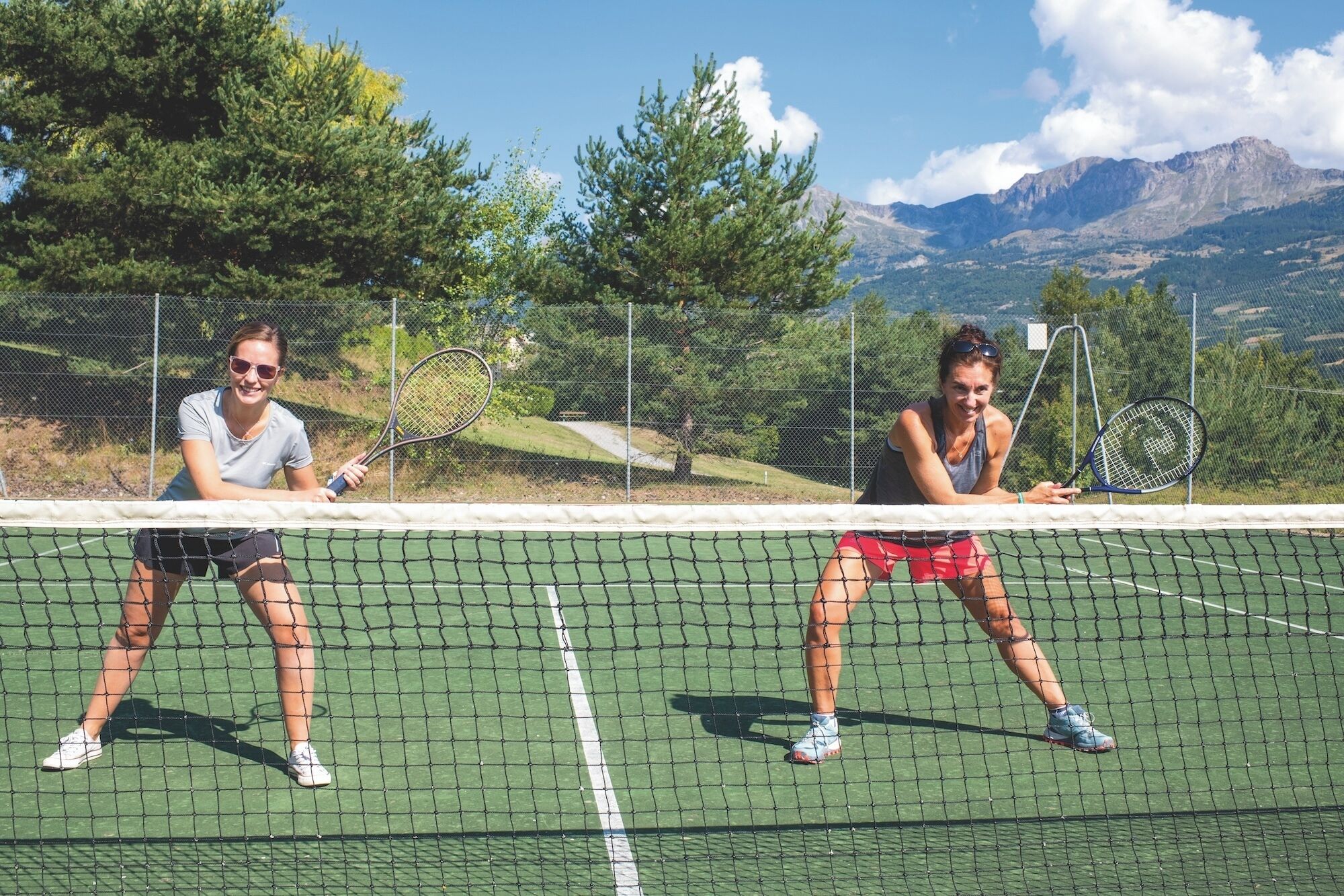 Two people playing tennis on a court with a net in the foreground and mountains in the background.