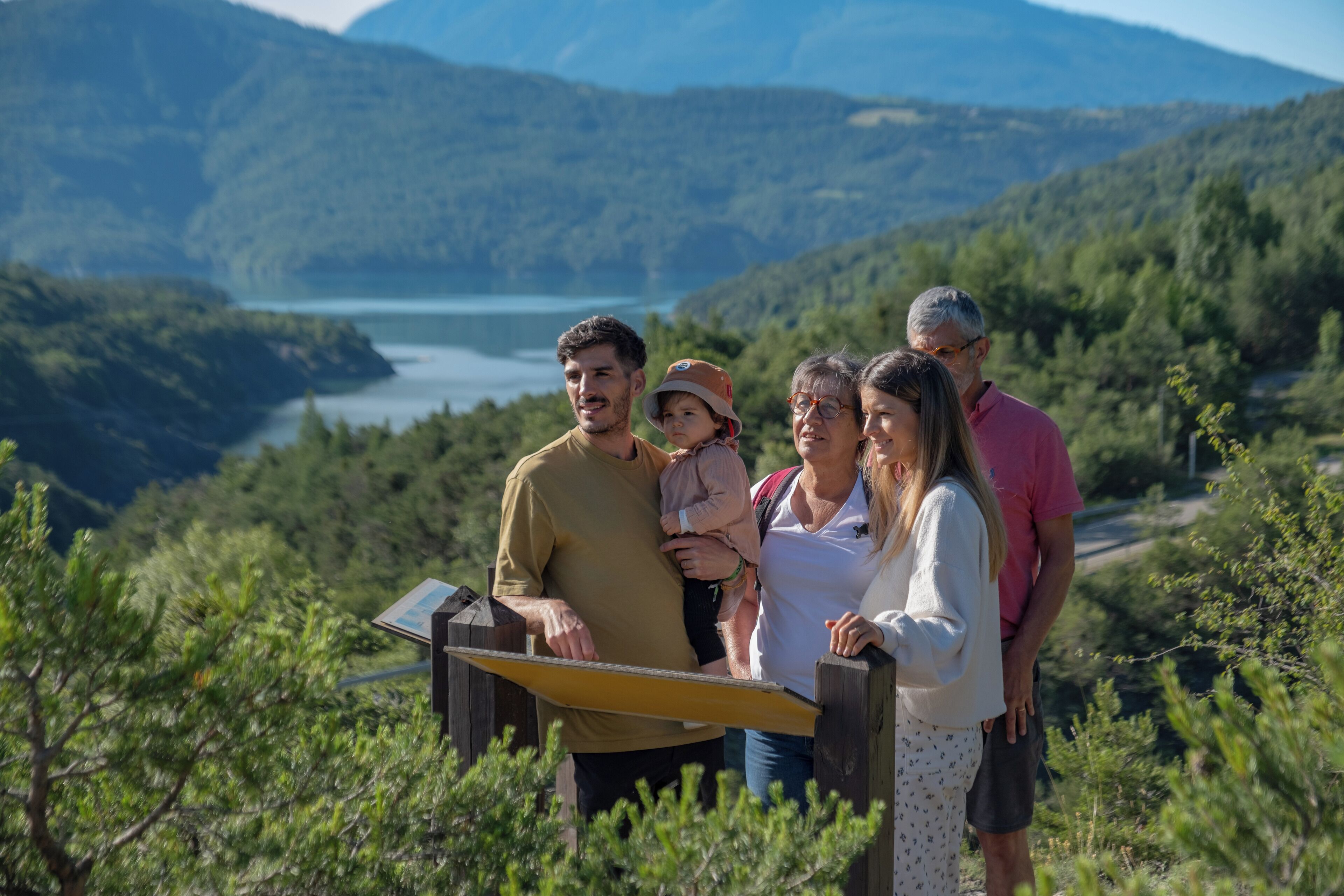 A family standing on a wooden viewing platform overlooking a lake and mountains.