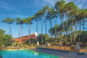 A swimming pool with lounge chairs and umbrellas, surrounded by tall trees and a building with a red roof. - Résidence Vacances Bleues Domaine de l'Agreou (Seignosse)